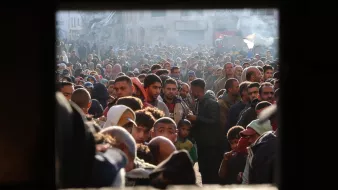 L'image montre une foule dense de personnes, probablement rassemblées dans un espace urbain. Les visages sont variés, et l'atmosphère semble chargée d'émotion et de tension. On peut voir des hommes, des femmes et des enfants, témoignant d'une situation de forte affluence, peut-être liée à un événement particulier comme une distribution d'aide ou une manifestation. Les lumières et l'arrière-plan laissent suggérer un contexte urbain avec des bâtiments visibles. L'image est encadrée par une ouverture, ce qui donne l'impression d'un regard extérieur sur ce rassemblement.